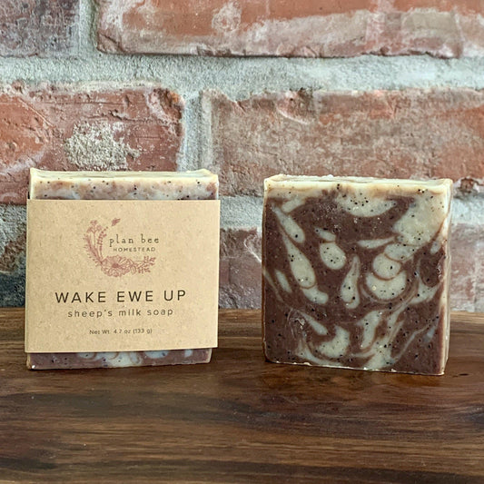 Brown and white swirled Wake Ewe Up sheep's milk soap with coffee grinds and cocoa powder on a wooded board with a brick background.